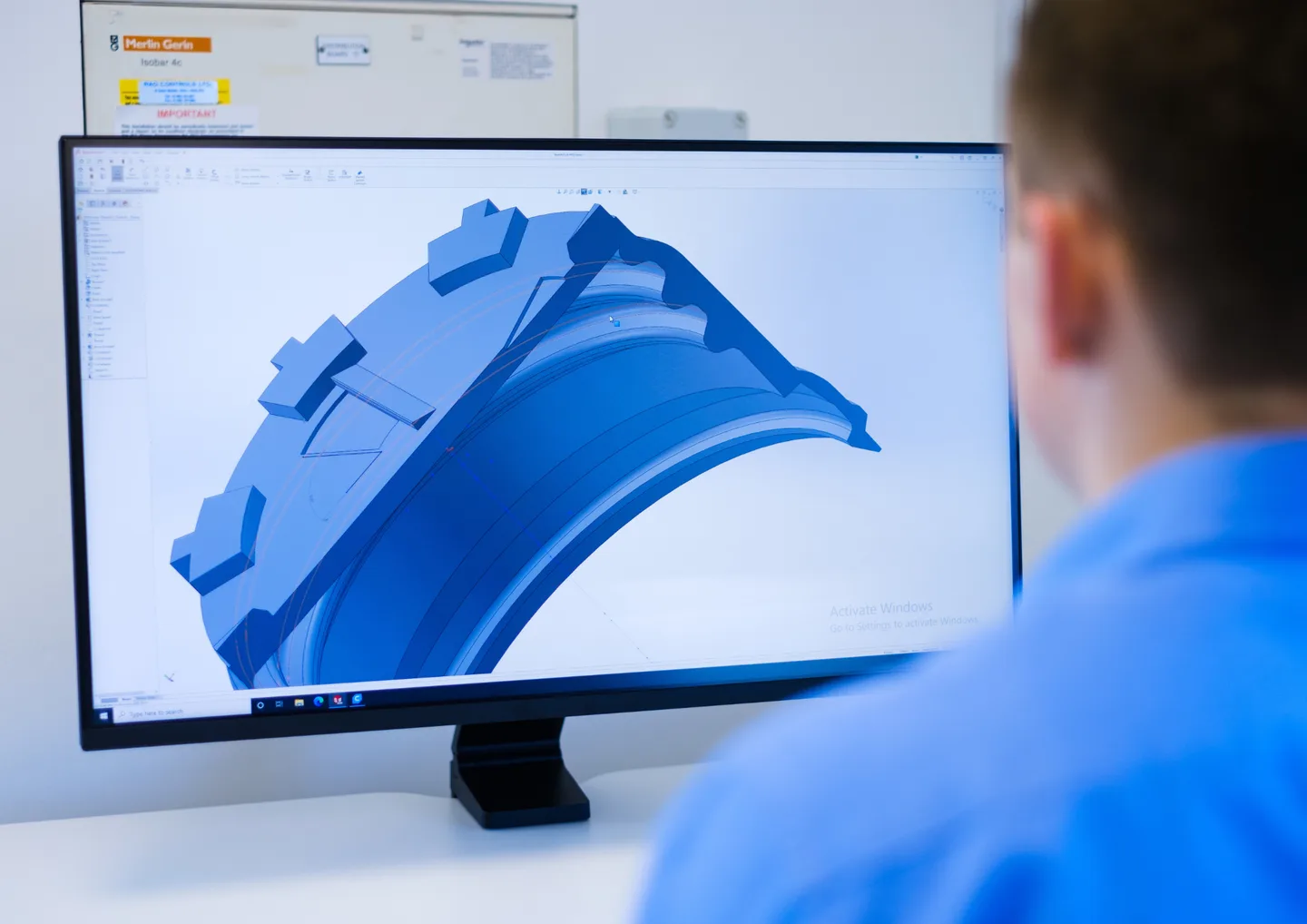 A young man in a pale blue shirt looks at a 3D render of a bottle cap on a computer screen.
